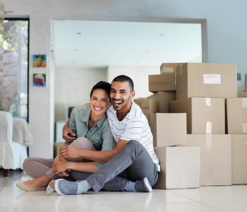 A young couple, a man and a woman, sit together on the floor of the living room in front of a stack of moving boxes